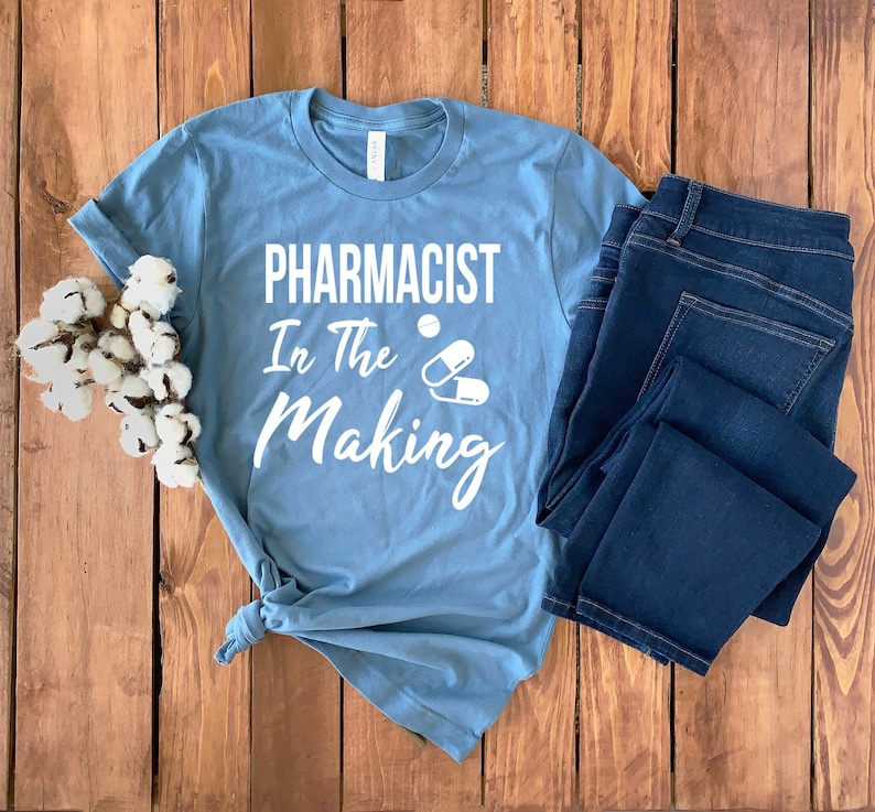 May include: A light blue t-shirt with the text "Pharmacist In The Making" in white lettering. The text is accompanied by a graphic of two white pills.