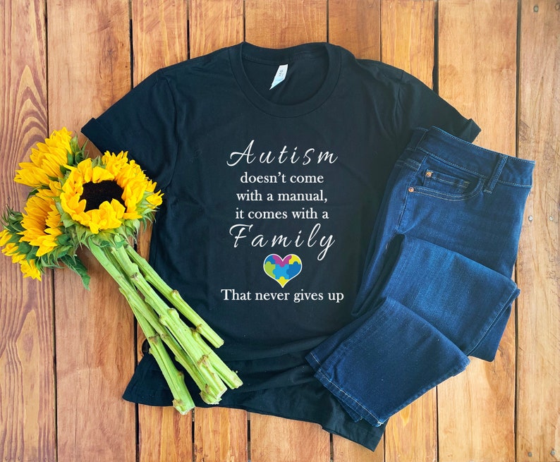 Op de afbeelding: Zwart T-shirt met witte tekst die luidt: "Autisme komt niet met een handleiding, het komt met een familie die nooit opgeeft". Een kleurrijk puzzelhart bevindt zich in het midden van het woord "Familie". Het shirt ligt op een houten oppervlak met een blauwe jeans en zonnebloemen.