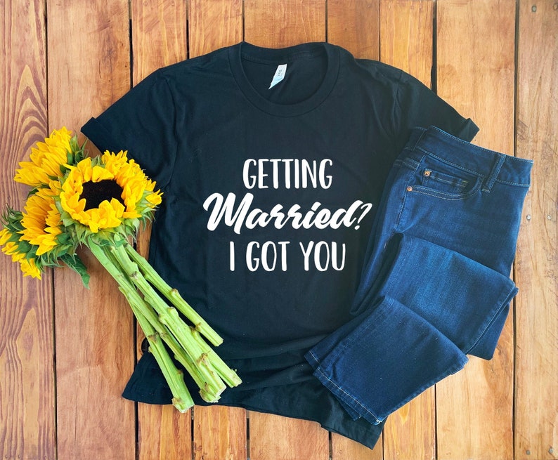 May include: Black t-shirt with white text that reads "Getting Married? I got you". The shirt is laid out on a wooden surface with a bouquet of sunflowers and a pair of blue jeans.