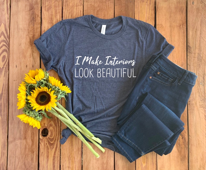 May include: A dark blue t-shirt with the text "I Make Interiors Look Beautiful" printed in white on the front. The t-shirt is laying on a wooden surface with a bouquet of sunflowers.
