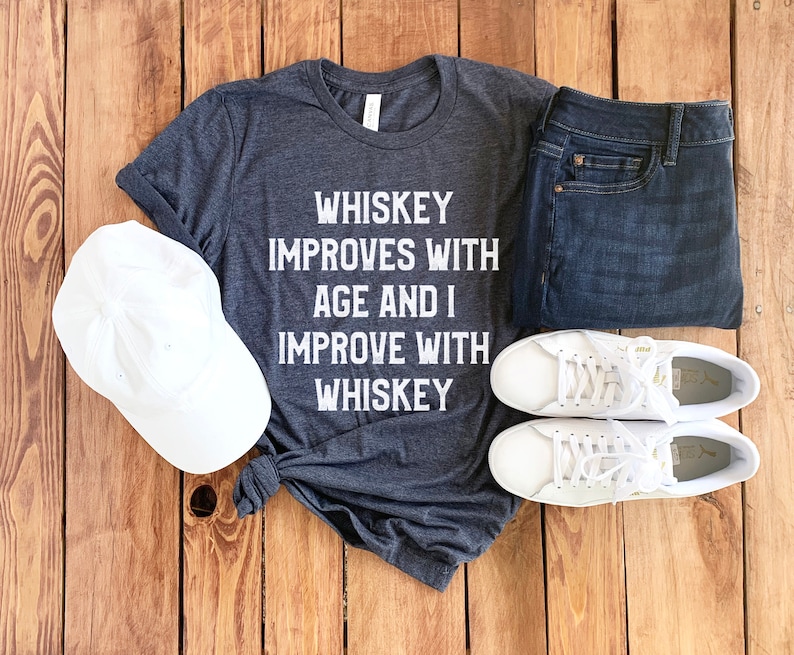 Peut inclure: Un t-shirt gris fonc&eacute; avec le texte "Whiskey improves with age and I improve with whiskey" imprim&eacute; en blanc. Le t-shirt est pos&eacute; sur une surface en bois avec une casquette de baseball blanche, un jean bleu et des baskets blanches.