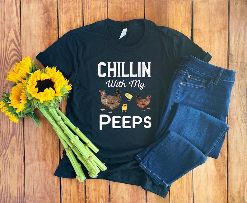 K&ouml;nnte beinhalten: Schwarzes T-Shirt mit wei&szlig;em Text "Chillin With My Peeps" und einer Grafik mit drei H&uuml;hnern. Das Shirt liegt auf einer Holzoberfl&auml;che mit einer blauen Jeans und einem Sonnenblumenstrau&szlig;.