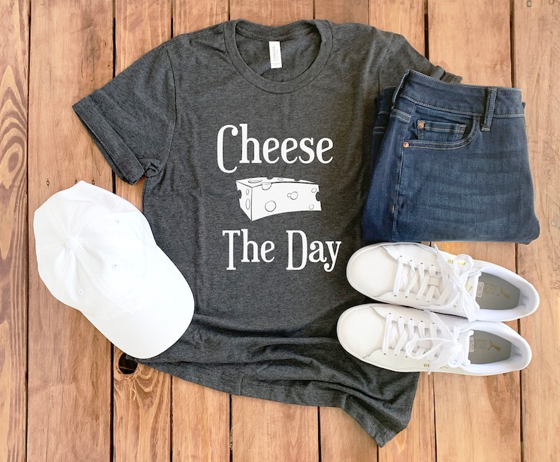 May include: A dark gray t-shirt with the text "Cheese The Day" and a graphic of a block of cheese. The shirt is paired with white sneakers, blue jeans, and a white baseball cap.
