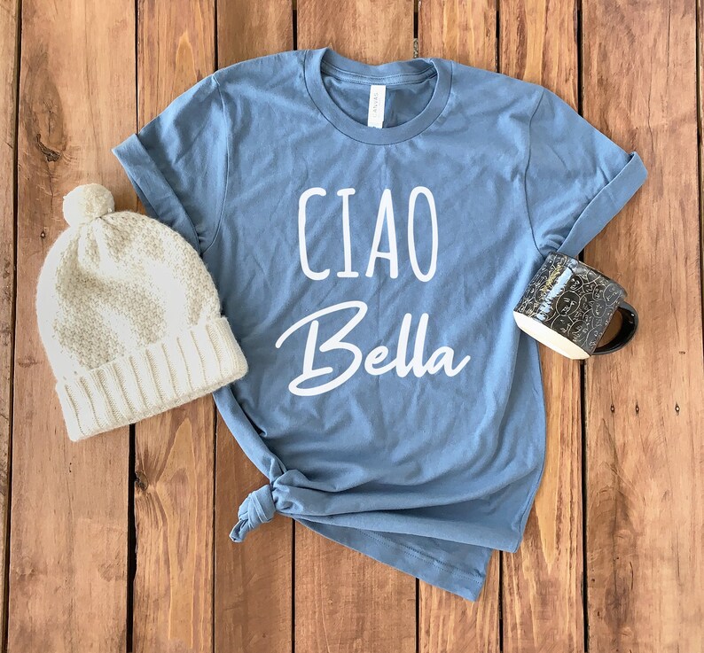 May include: A blue t-shirt with the text "Ciao Bella" in white lettering. The shirt is on a wooden surface with a beige knitted hat and a black mug.