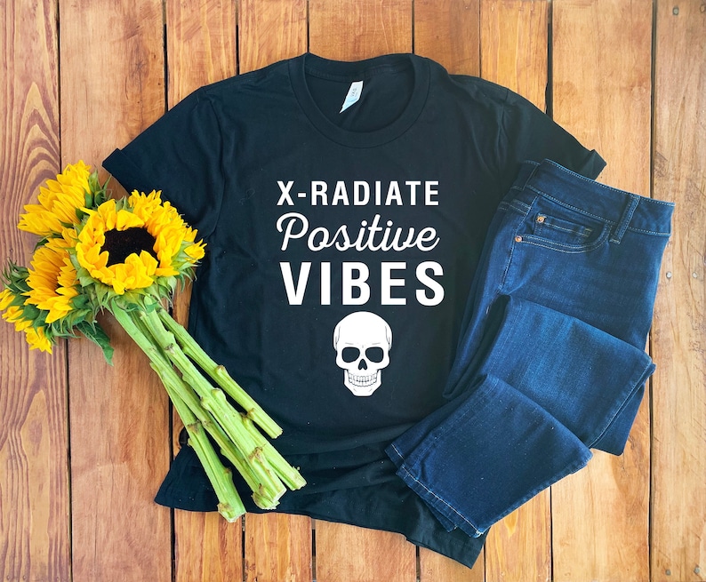 Op de afbeelding: Zwart T-shirt met witte tekst die "X-RADIATE Positive VIBES" zegt en een witte schedel afbeelding. Het shirt ligt op een houten oppervlak met een blauwe jeans.
