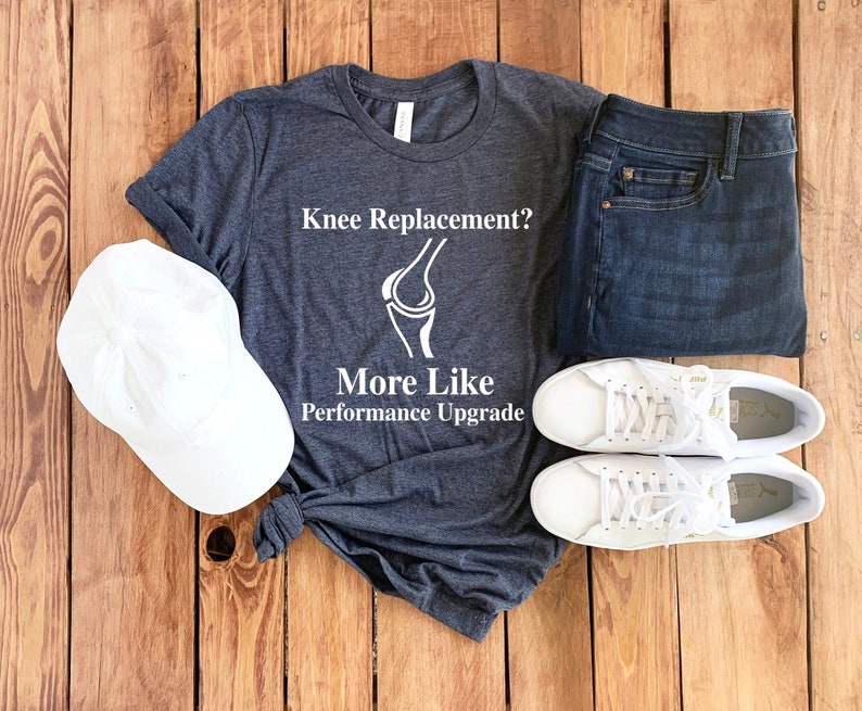 May include: A dark gray t-shirt with white text that reads "Knee Replacement? More Like Performance Upgrade". The shirt features a white outline of a knee joint. The shirt is laid out on a wooden surface with a white baseball cap, a pair of blue jeans, and a pair of white sneakers.