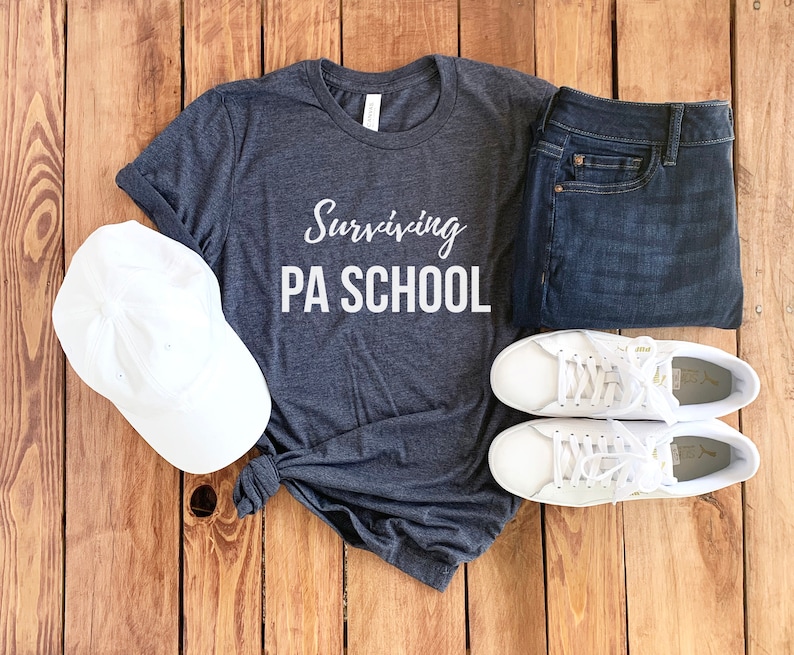 Op de afbeelding: Een donkergrijs T-shirt met de tekst "Surviving PA School" in witte letters. Het shirt ligt op een houten oppervlak met een witte baseballpet, een blauwe spijkerbroek en twee witte sneakers.