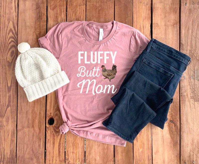 May include: A pink t-shirt with the text "FLUFFY Butt Mom" and a graphic of a chicken. The shirt is laid out on a wooden surface with a white beanie and blue jeans.