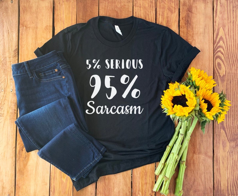 Pu&ograve; includere: Maglietta nera con testo bianco che recita "5% Serio 95% Sarcasmo". La maglietta &egrave; appoggiata su una superficie di legno con un paio di jeans blu e un mazzo di girasoli.