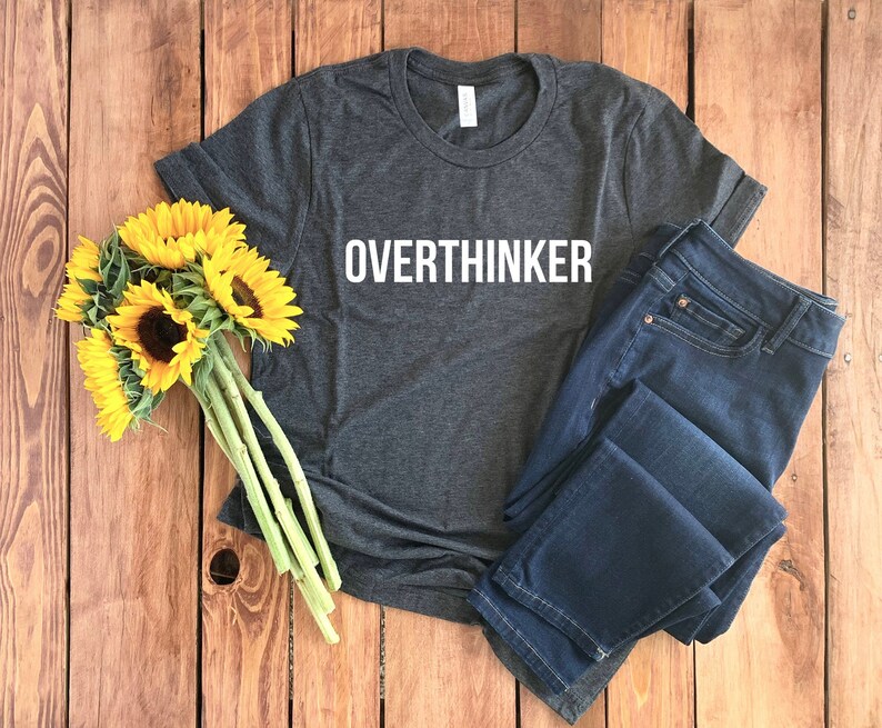 May include: A dark gray t-shirt with the word "Overthinker" printed in white on the front. The shirt is laid flat on a wooden surface with a bouquet of sunflowers and a pair of blue jeans.