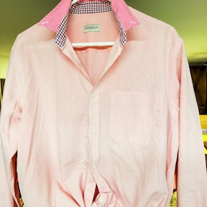 May include: A long-sleeved button-down shirt in a light pink color. The shirt features a collar with a pink and white gingham pattern on the outer side and a dark blue and white checkered pattern on the inner side. A label reads Portobellos.