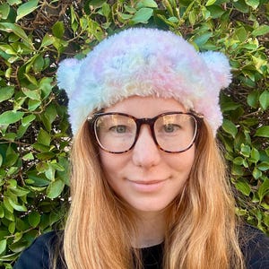 May include: A woman with long, wavy, light brown hair is wearing a fuzzy, pastel pink, blue, and white tie-dye beanie with ear flaps. She is wearing brown-rimmed glasses and a black shirt.