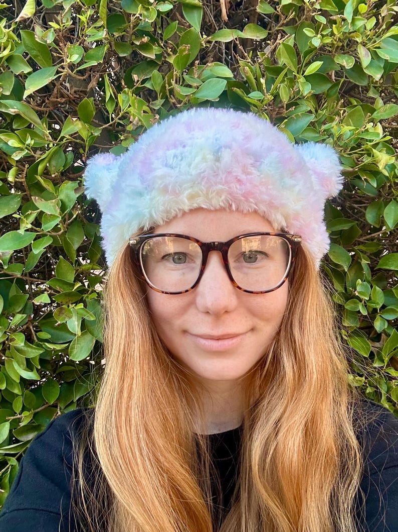 May include: A woman with long, wavy, light brown hair is wearing a fuzzy, pastel pink, blue, and white tie-dye beanie with ear flaps. She is wearing brown-rimmed glasses and a black shirt.