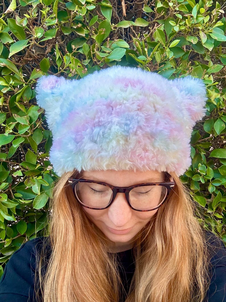 May include: A fluffy pastel tie-dye beanie with fuzzy ear flaps. The beanie is made of soft, plush fabric and has a light blue, pink, and yellow color scheme.