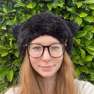 May include: A black fuzzy hat with cat ears. The hat is worn by a person with long, red hair and glasses.