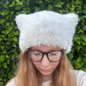 May include: A white fuzzy hat with cat ears. The hat is made of a soft, plush material and has a simple design.