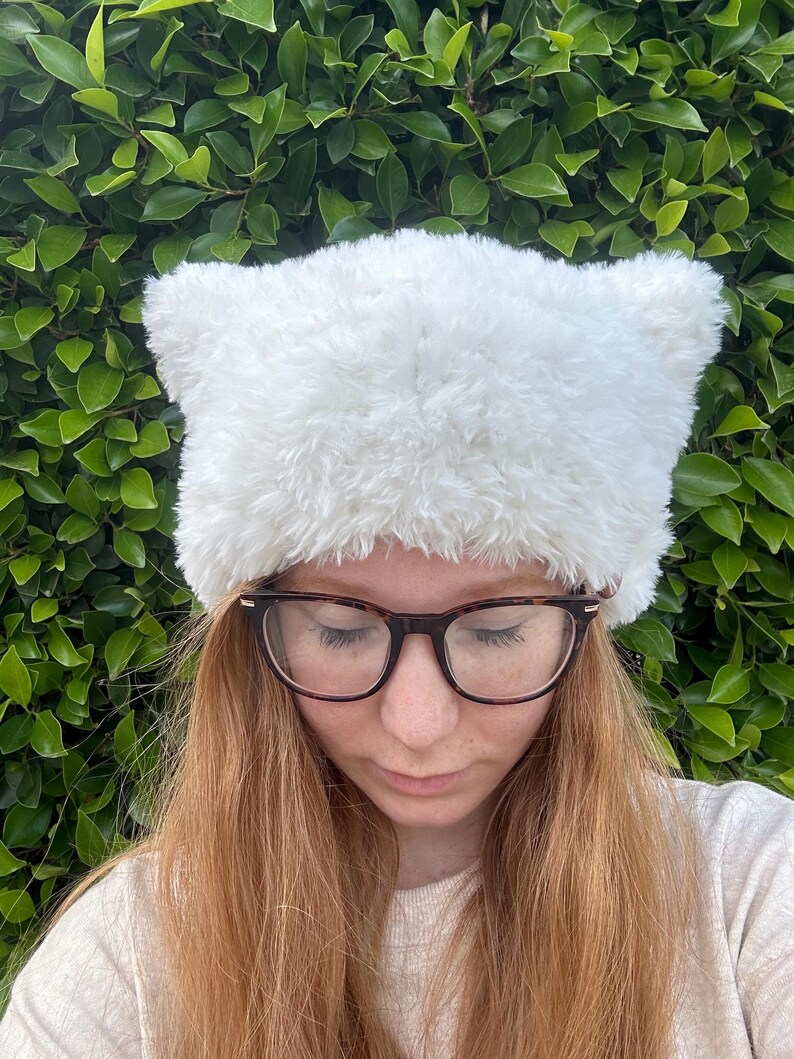 May include: A white fuzzy hat with cat ears. The hat is made of a soft, plush material and has a simple design.