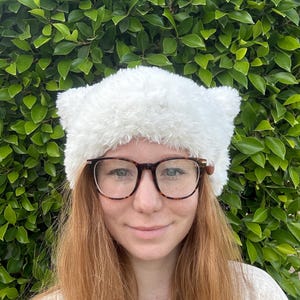 May include: A white fuzzy cat ear headband with a fluffy texture. The headband is worn by a person with long, red hair and glasses.