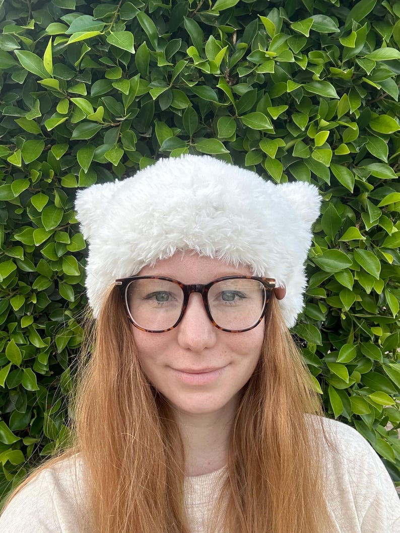 May include: A white fuzzy cat ear headband with a fluffy texture. The headband is worn by a person with long, red hair and glasses.