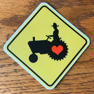 May include: A yellow diamond-shaped road sign with a black silhouette of a tractor and a farmer driving it. The tractor has a red heart on the back tire.
