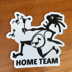 May include: Black and white sticker of a cartoon figure playing drums with the text "HOME TEAM" below.