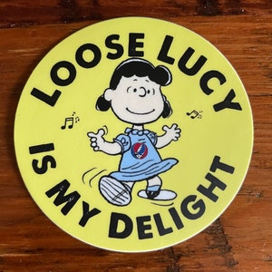 May include: A yellow circle sticker with the text "Loose Lucy is my delight" in black lettering. A cartoon image of Lucy from the Peanuts comic strip is in the center of the sticker. Lucy is wearing a blue dress with a Grateful Dead logo on it and is dancing with her arms outstretched.