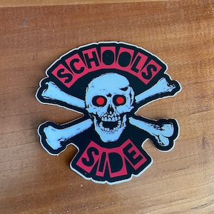 May include: A sticker featuring a skull with red eyes and crossed bones. The words "SCHOOL'S SIDE" are in red, outlined in black, and set against a white background.