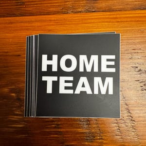 May include: A stack of black square stickers with the words "HOME TEAM" in bold white letters. The stickers are on a wooden surface with a natural wood grain pattern.