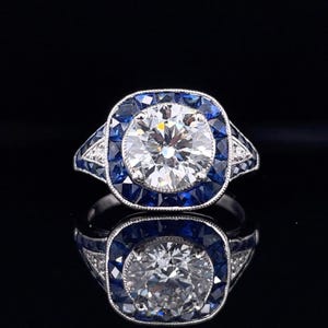 A professional 1.90ct round brilliant lab-grown diamond ring in a platinum Art Deco setting. The center stone is encircled by a geometric double halo of French-cut blue sapphires and pavé-set white and blue diamonds. Featuring milgrain edges and an antique target-style design on a polished band.