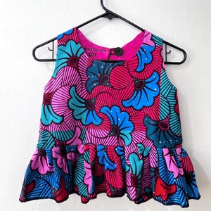 May include: A sleeveless peplum top featuring a vibrant floral pattern in shades of hot pink, bright blue, and teal. The garment has a round neckline, a single black button closure at the back, and a ruffled hem. The interior label reads Dzwa Designs 10Y.