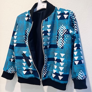 May include: A blue bomber jacket with a geometric pattern of white triangles and lines. The jacket has a black collar, cuffs, and hem, and a silver zipper. The jacket is hanging on a white hanger.