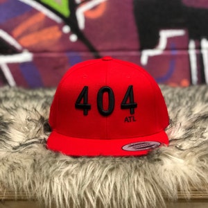 May include: A red baseball cap with a black 404 ATL logo. The cap is on a fuzzy red surface.