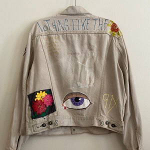May include: A beige denim jacket with various painted designs, including a large eye, flowers, and the text "NOTHING LIKE THE" and "91X".