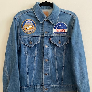 May include: A blue denim jacket with two NASA patches. The left patch features a space shuttle and the text "Dryden Flight Research Facility". The right patch features a star and the text "NASA Ames Research Center".
