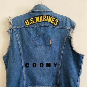 Peut inclure: Un gilet en jean bleu avec des bords effilochés et un patch jaune "U.S. Marines" au dos. Le gilet porte une inscription noire "COONY" sur le devant.