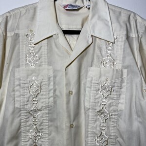 May include: A white short-sleeved button-down shirt with embroidered details. The shirt has a Cuban collar and a label that reads "Ronani" on the front.