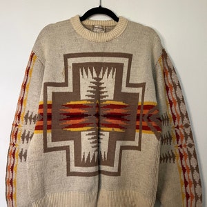 Puede incluir: Un suéter de color crema con un patrón geométrico marrón, naranja y rojo. El suéter tiene cuello redondo y mangas largas.