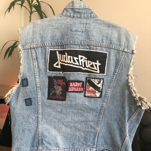 Puede incluir: Un chaleco vaquero azul claro con varios parches, incluyendo un parche grande de "Judas Priest", un parche de "Randy Rhoads" y un parche de "Led Zeppelin". El chaleco tiene bordes deshilachados y está desgastado.