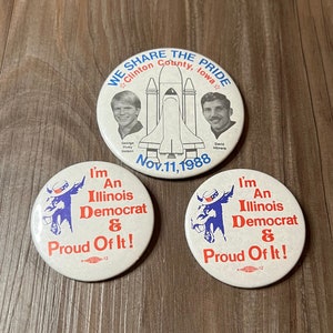 May include: Three round buttons with political campaign slogans. The top button features a space shuttle and the text "We Share The Pride Clinton County, Iowa Nov. 11, 1988". The two bottom buttons feature a cartoon donkey and the text "I'm An Illinois Democrat & Proud Of It!"