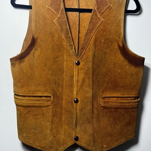 Może przedstawiać: Brązowa kamizelka zamszowa z wyciętym kołnierzem, trzema guzikami i dwiema kieszeniami z przodu.