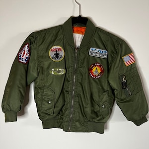 Könnte beinhalten: Eine grüne Bomberjacke mit mehreren Patches, darunter "Apache", "F-16 Fighter Pilot" und "F-22 Raptor".