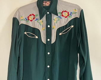 May include: A green long-sleeved shirt with a white collar and buttons. The shirt has a grey yoke with colourful floral embroidery. The cuffs also have floral embroidery.
