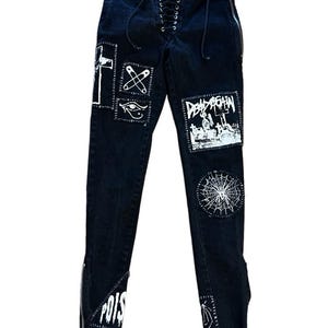 May include: Black denim jeans with multiple white patches featuring various designs, including a cross, an eye, and a spiderweb. The jeans have a lace-up front, zippers on the sides, and the word "POIS" on one leg.
