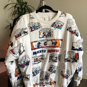 May include: White long-sleeve sweatshirt with a print of Mickey Mouse comic strips. The print features Mickey Mouse in various scenes and includes the text "Mickey Mouse".