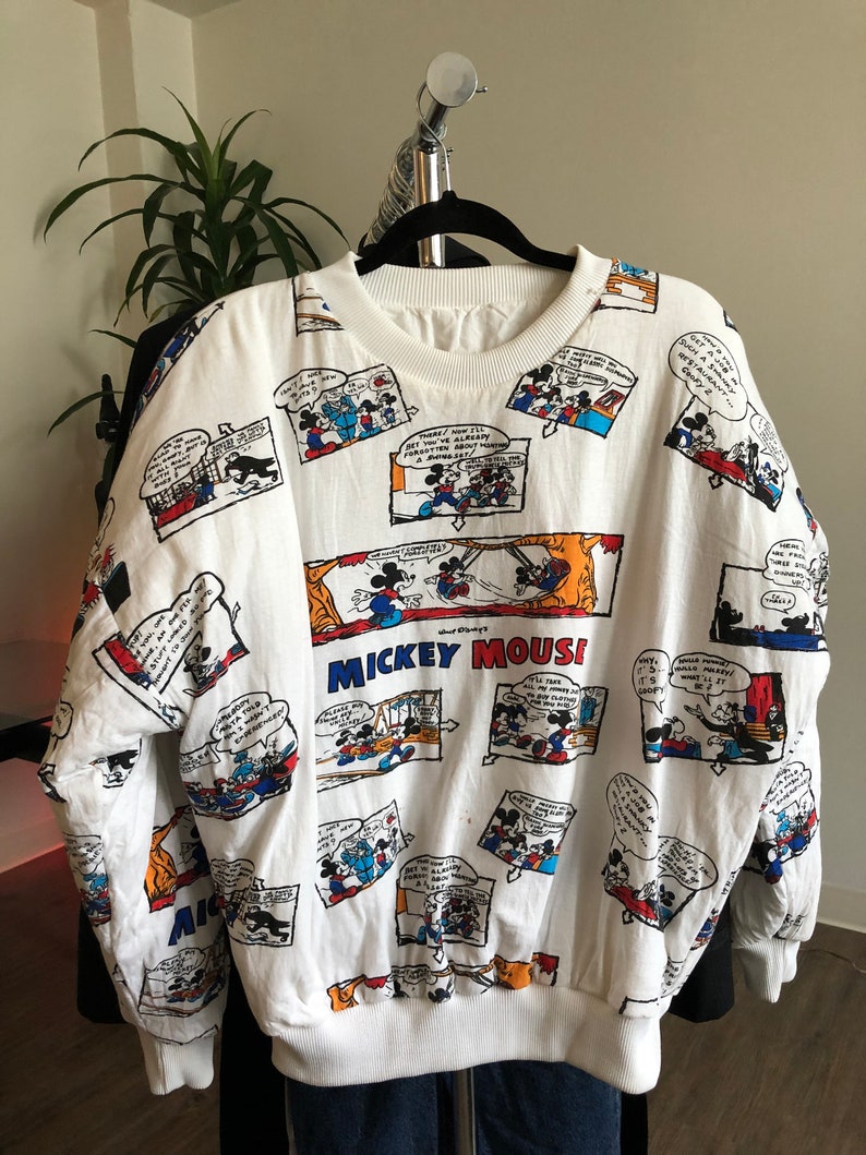 May include: White long-sleeve sweatshirt with a print of Mickey Mouse comic strips. The print features Mickey Mouse in various scenes and includes the text "Mickey Mouse".
