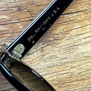 K&ouml;nnte beinhalten: Nahaufnahme eines schwarzen Sonnenbrillenb&uuml;gels mit dem wei&szlig;en Aufdruck "B&L RAY-BAN U.S.A.". Der Sonnenbrillenb&uuml;gel ist gl&auml;nzend und reflektiert das Licht. Das Scharnier ist sichtbar, und der Hintergrund ist eine Holzoberfl&auml;che.
