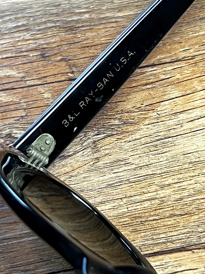 K&ouml;nnte beinhalten: Nahaufnahme eines schwarzen Sonnenbrillenb&uuml;gels mit dem wei&szlig;en Aufdruck "B&L RAY-BAN U.S.A.". Der Sonnenbrillenb&uuml;gel ist gl&auml;nzend und reflektiert das Licht. Das Scharnier ist sichtbar, und der Hintergrund ist eine Holzoberfl&auml;che.