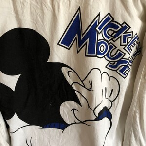May include: White long-sleeve shirt with a black and white Mickey Mouse graphic and blue text that says "Mickey Mouse".