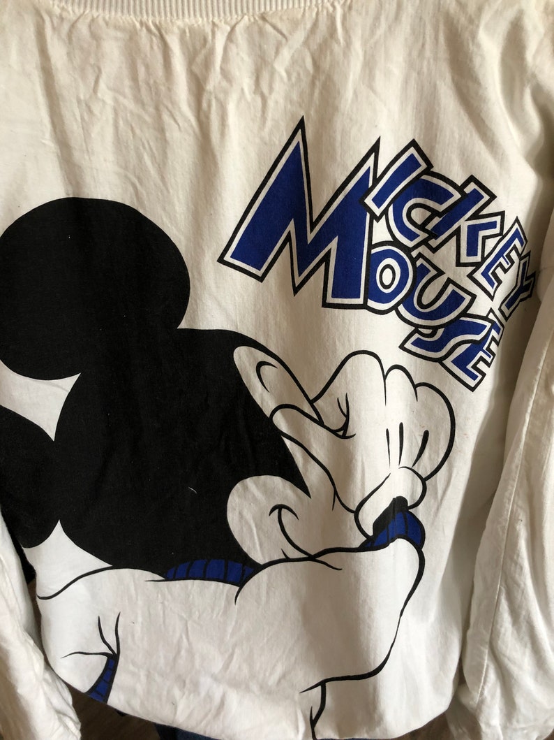 May include: White long-sleeve shirt with a black and white Mickey Mouse graphic and blue text that says "Mickey Mouse".
