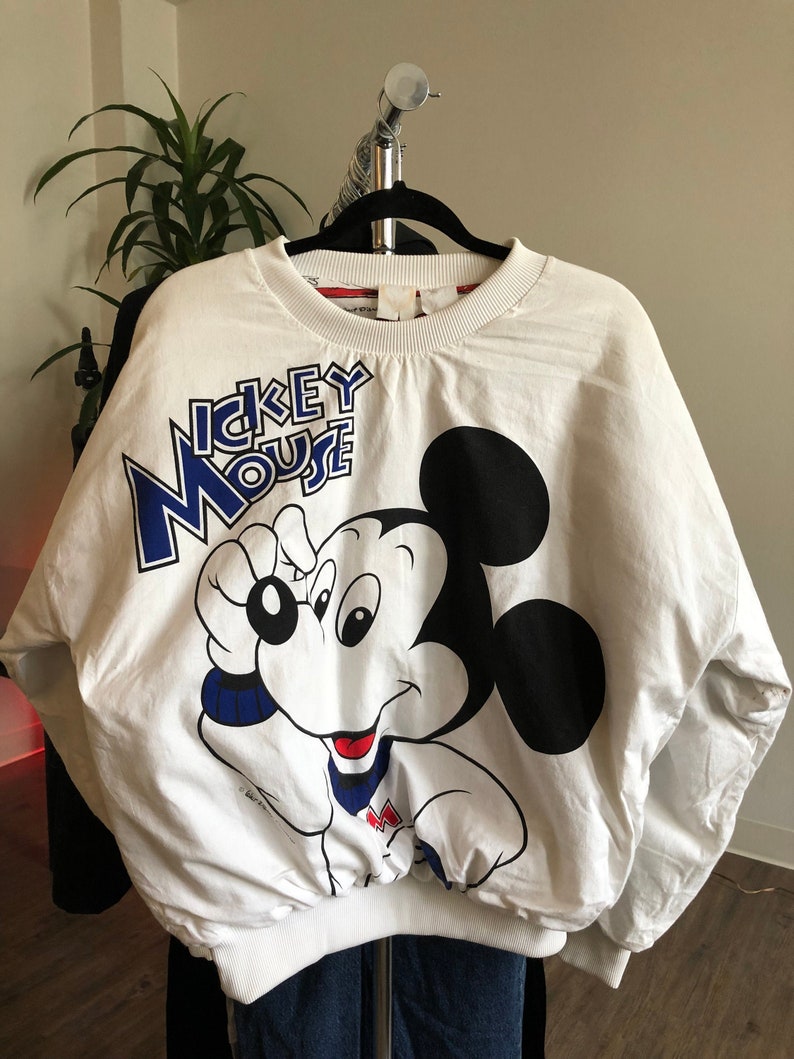 May include: White sweatshirt with a large Mickey Mouse graphic on the front. The graphic is in blue, black, and red. The text "Mickey Mouse" is printed in blue above the graphic.